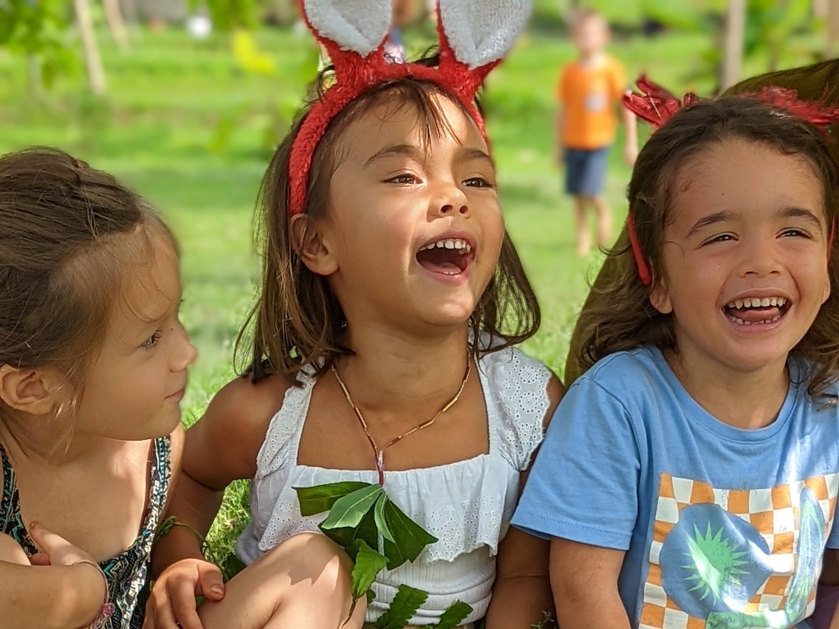Three children sit together outdoors, two laughing and one wearing a red headband with ears.