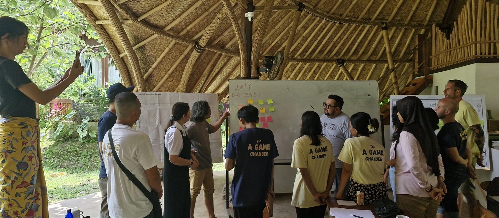 Adults gather around a whiteboard in an open bamboo pavilion.
