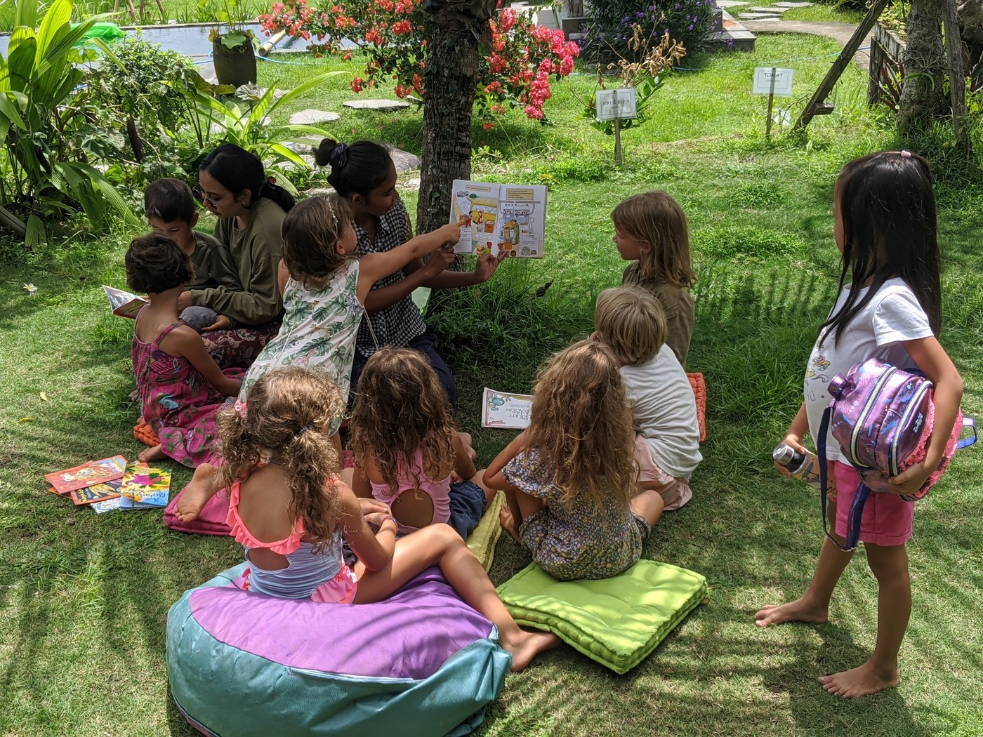 An adult holds an open book while children sit on cushions in a garden.