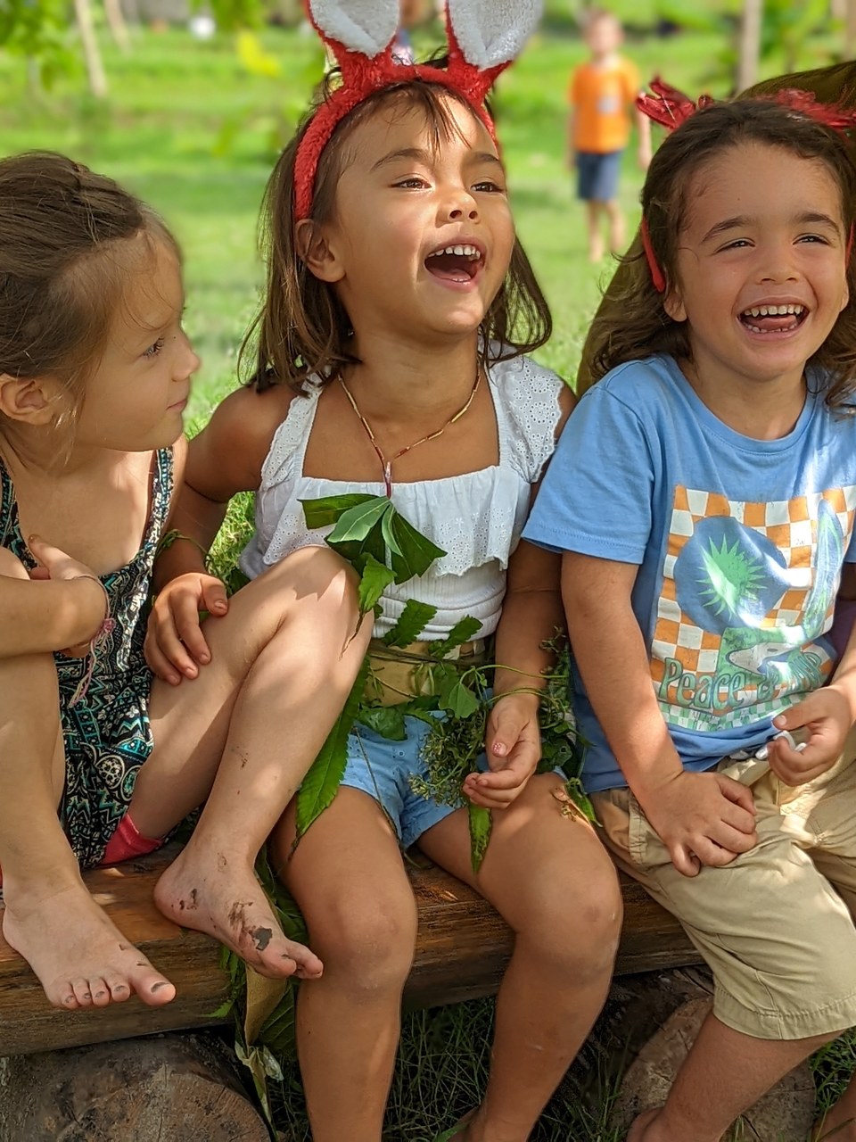 Three children sit together outdoors, two laughing and one wearing a red headband with ears.